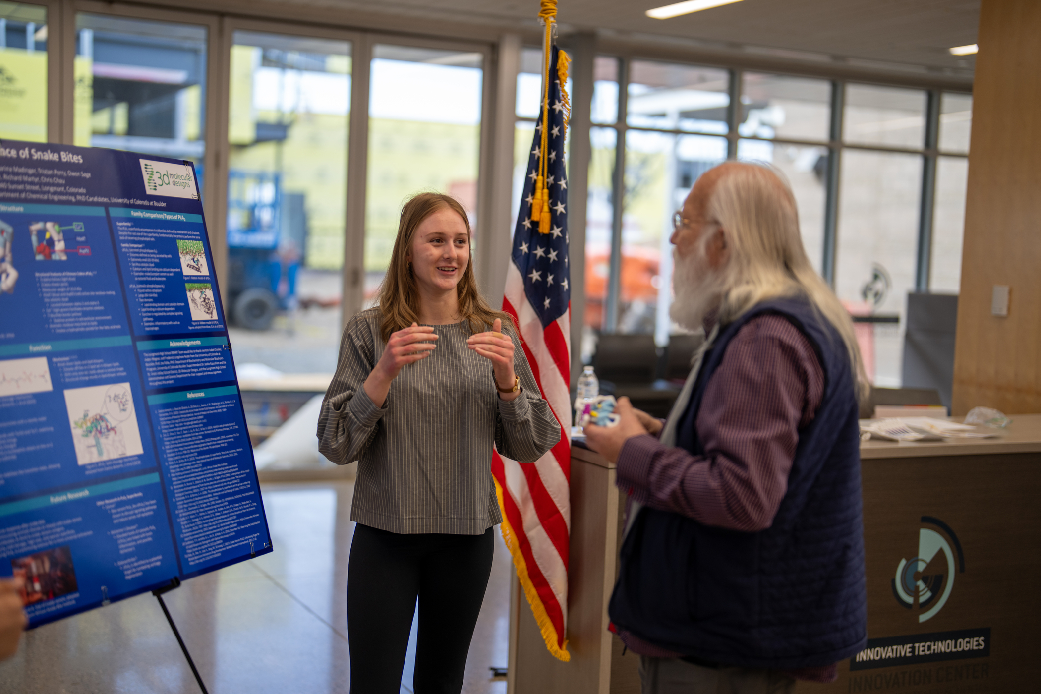 A student presents research on snake bites to an older man, with a poster and an American flag in the background.
