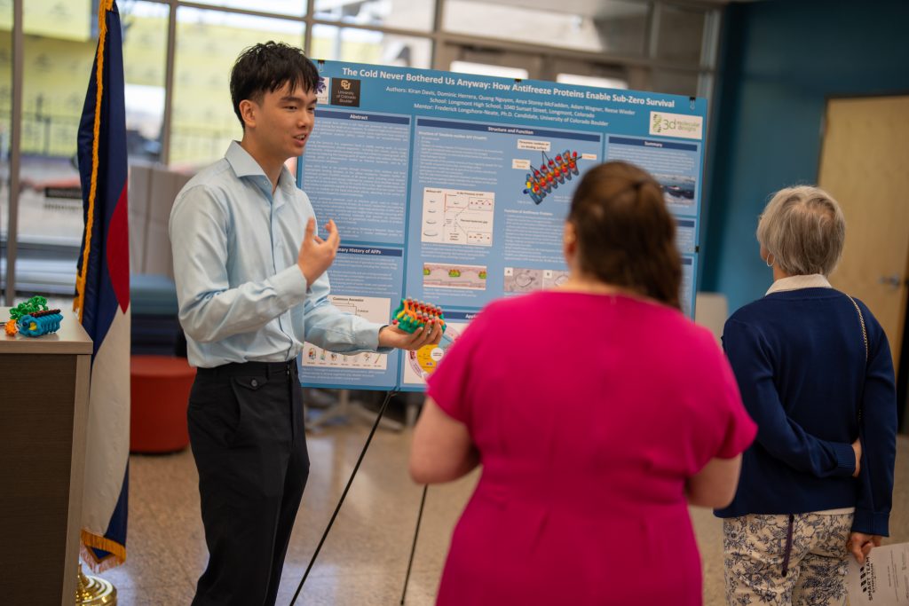 A young presenter explains his research on antifreeze proteins to two attendees at a science event.