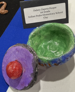 Colorful clay art piece featuring a green bowl and a purple lid with a red knob, labeled with the artist's details.
