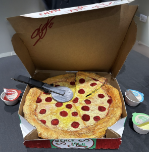 A pizza replica in a box, with pepperoni, a pizza cutter, and dipping sauce containers beside it.
