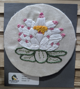 Embroidery of a white lotus flower with pink tips and a yellow center, mounted on a dark backing.