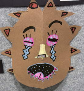 A colorful, spiky mask made from brown cardboard, featuring exaggerated facial features and vibrant designs.