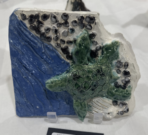Ceramic artwork depicting a green turtle on a textured blue and white background with black pebbles.