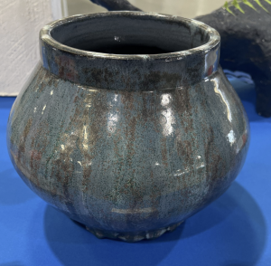A round, textured ceramic vase with a glossy dark blue-green finish, placed on a blue surface.