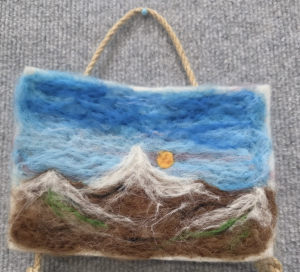 A felt artwork depicting mountains under a blue sky with a sun, hanging by a twine loop.
