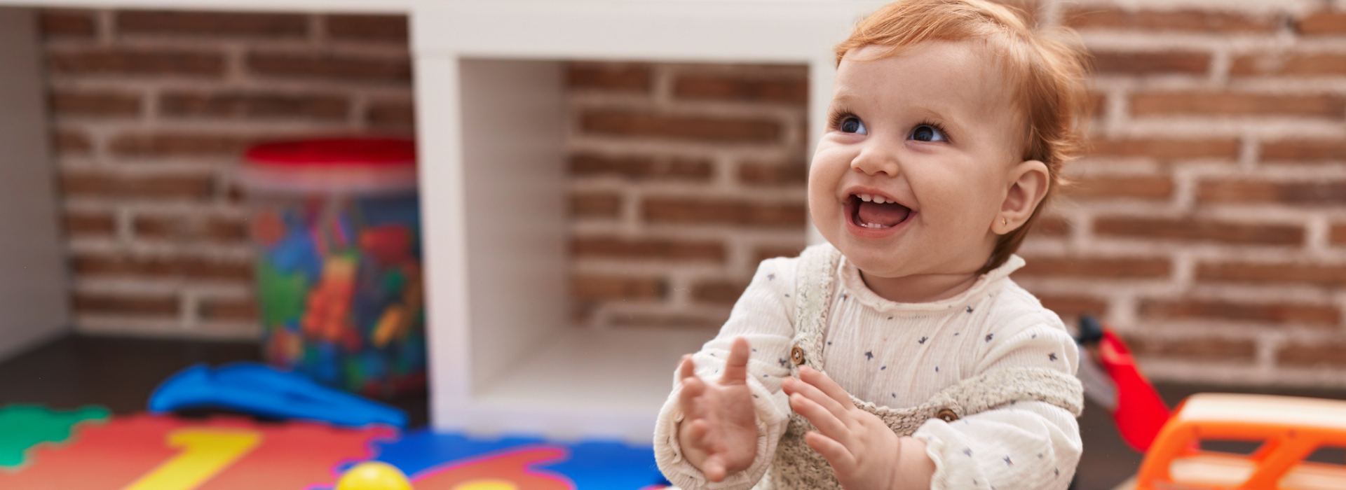 A smiling baby with red hair claps hands while sitting on a colorful play mat in a cozy room.