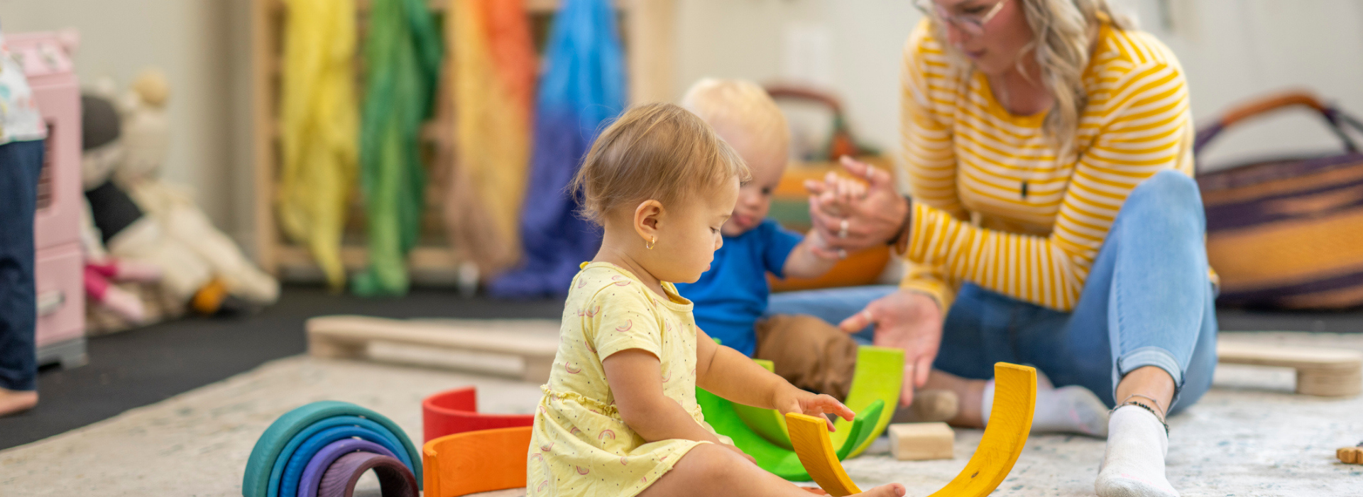 A child plays with colorful toys on a mat while an adult engages with another child nearby in a bright room.