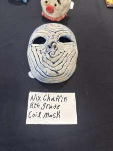 A ceramic mask with textured lines, displayed on a black table, labeled as made by an 8th grader named Nix Chaffin.