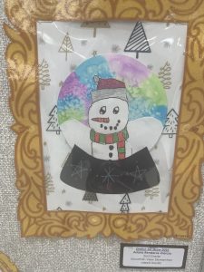 A colorful drawing of a snowman in a decorative frame, displayed at a school art show.