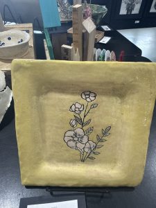 A square, yellow ceramic plate features a delicate floral design with pink flowers and green leaves.