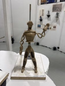 A clay sculpture of a humanoid figure with articulated limbs, displayed on a wooden base in an art exhibit.