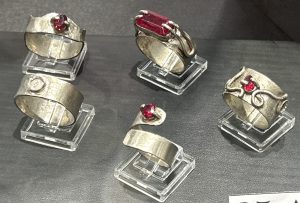 Five silver rings with red gemstones displayed on clear stands against a dark background.