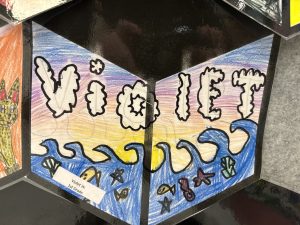 Colorful artwork featuring the name "Violet" with ocean waves, fish, and a sunset background. Created by a first grader.