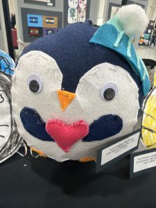 A colorful, felt penguin sculpture with big eyes, a heart-shaped belly, and a teal hat, displayed at an art exhibit.