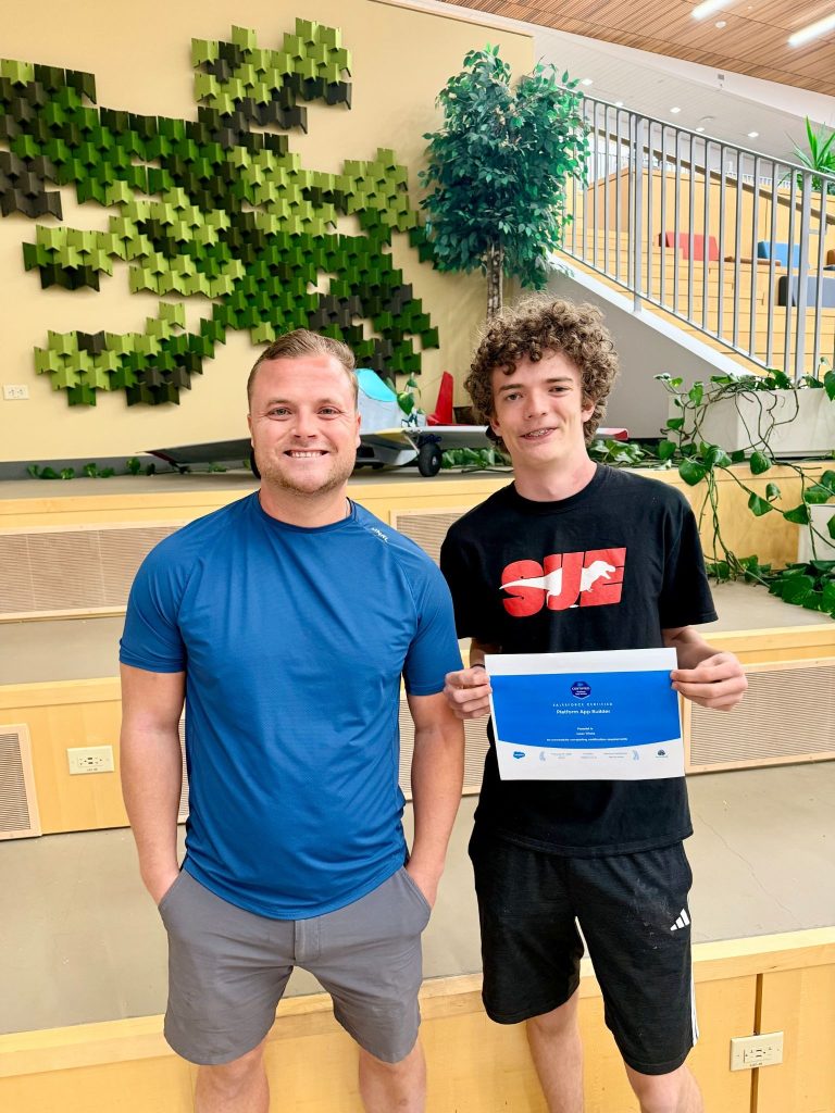Dos jóvenes posan sonrientes; uno de ellos sostiene un certificado, con un diseño de pared verde y plantas al fondo.