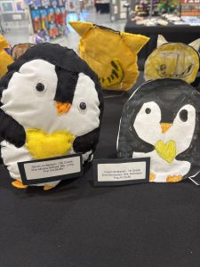 Two penguin art pieces are displayed: a plush penguin and a drawing, both labeled with students' names and grades.