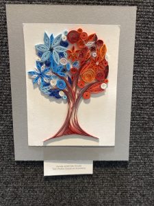 Colorful quilled paper artwork depicting a tree with flowers in red, blue, and white, mounted on a gray background.