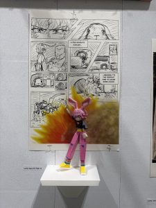 A colorful paper figure stands on a shelf beside a comic strip with various panels and speech bubbles.