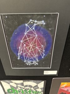 A colorful drawing of a constellation against a dark, swirling background, framed and displayed on a wall.
