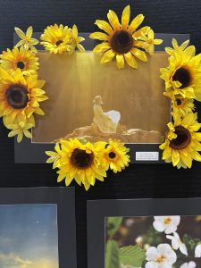 A framed photo of a woman in a white dress surrounded by artificial sunflowers, displayed on a black background.