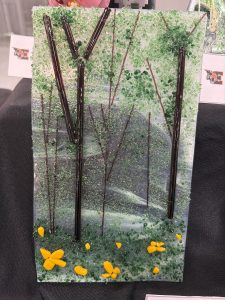A glass artwork depicting a forest scene with green foliage, dark branches, and yellow flowers at the base.