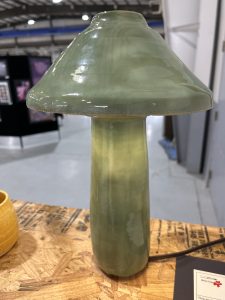A green ceramic lamp with a mushroom shape, placed on a wooden surface, with art displays in the background.