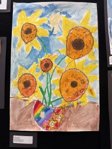 A colorful child’s drawing of sunflowers in a vase, with a blue sky background and a rainbow-colored pot.