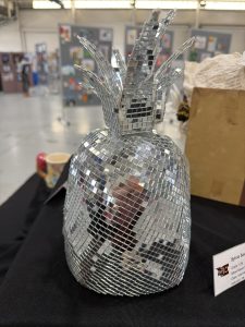 A reflective, mosaic pineapple sculpture sits on a black table, with art displays in the background.