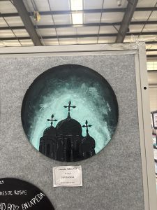 Cuadro circular en el que se representan cúpulas oscuras con cruces sobre un fondo verde azulado y negro, expuesto sobre un tablero gris.
