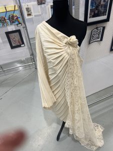 A cream-colored garment on a mannequin, featuring pleats and a lace overlay, displayed among various artworks.