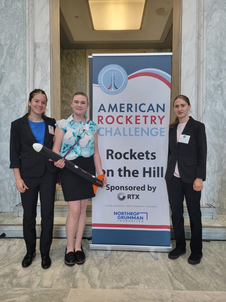 Three young women in business attire pose with a model rocket beside a banner for the American Rocketry Challenge.