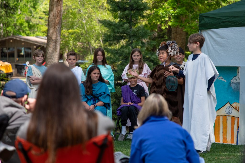 Children in costumes perform a play outdoors, with an audience seated on the grass and trees in the background.