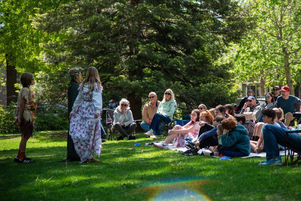 Children perform on stage in a park while an audience of families watches from the grass in a sunny setting.