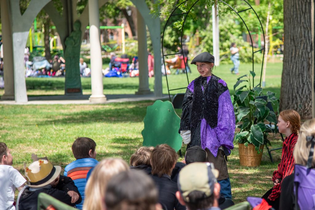A child in a purple costume performs on stage while an audience of children watches in a park setting.