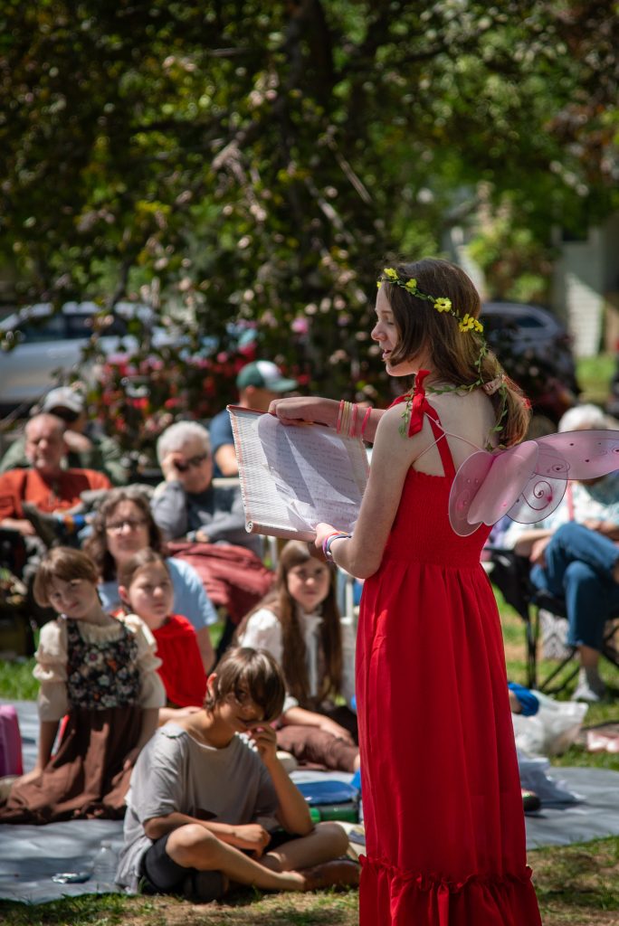 A girl in a red dress with fairy wings reads from a book to an audience seated on the grass in a park.