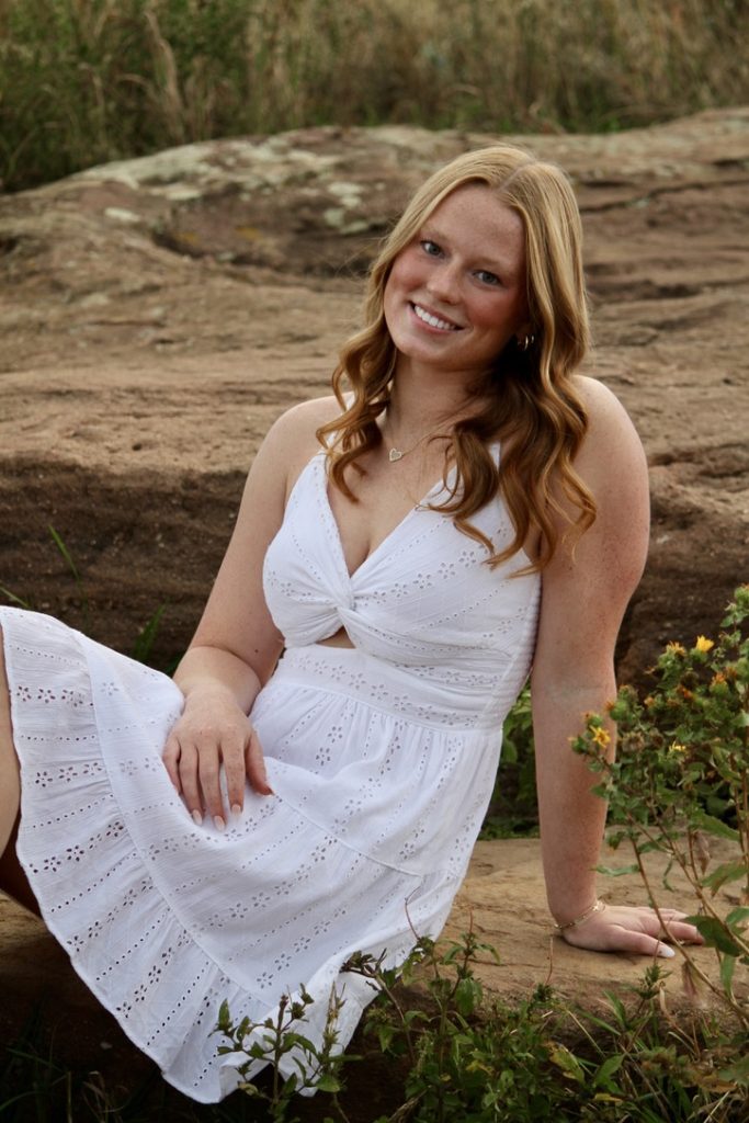 A smiling person with long wavy hair sits on a rock, wearing a white dress surrounded by greenery and flowers.