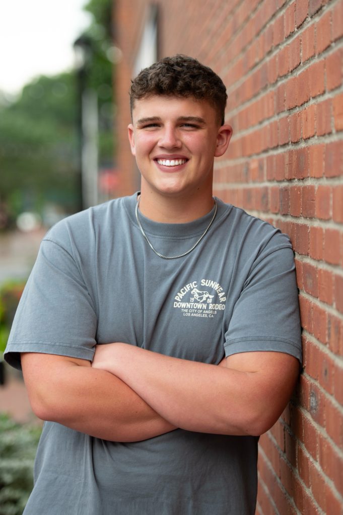 Young man in a gray t-shirt smiles confidently with arms crossed against a brick wall in an outdoor setting.