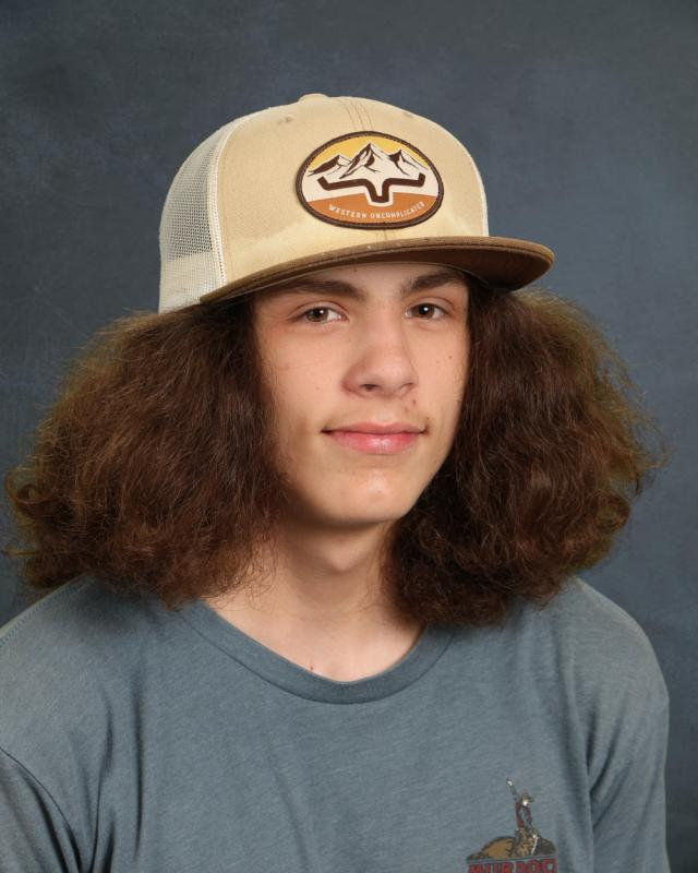 Teen with curly hair wearing a beige cap and gray t-shirt, smiling against a blue background.