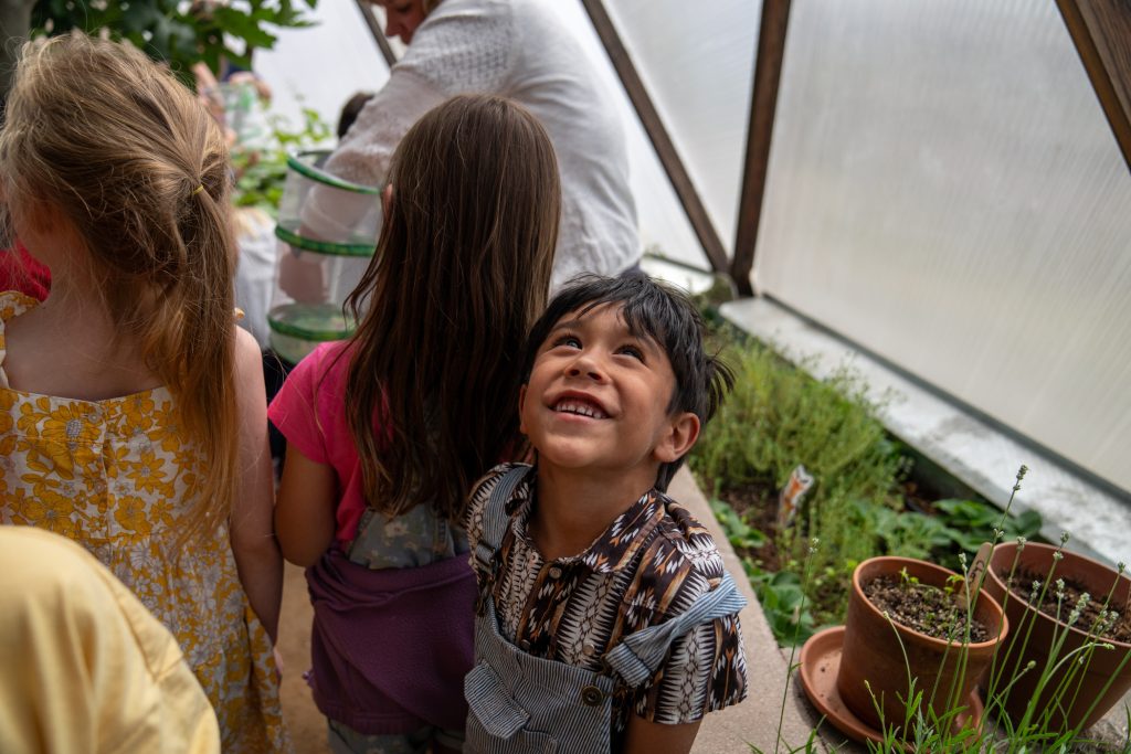 Un niño sonriente mira hacia arriba rodeado de otros niños en un invernadero lleno de plantas.