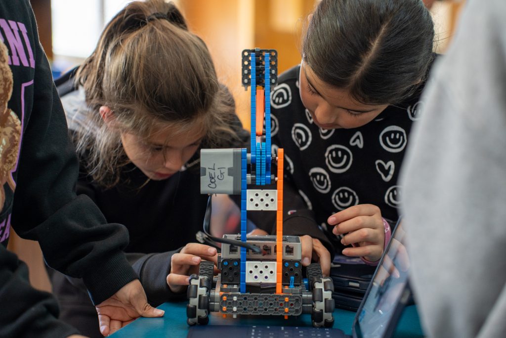 Two children focus intently on building a robot with colorful blocks on a table, surrounded by classmates.