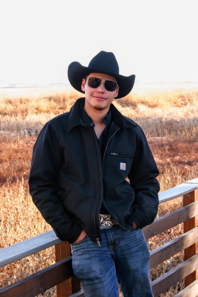 A young man in a black cowboy hat and sunglasses stands confidently by a wooden railing, with a grassy landscape behind him.