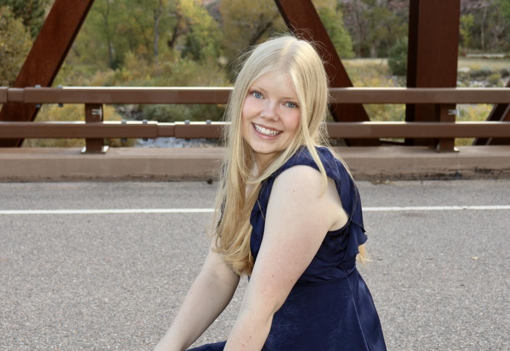 A smiling young woman with long blonde hair sits on a road, wearing a navy dress, with trees and a bridge in the background.
