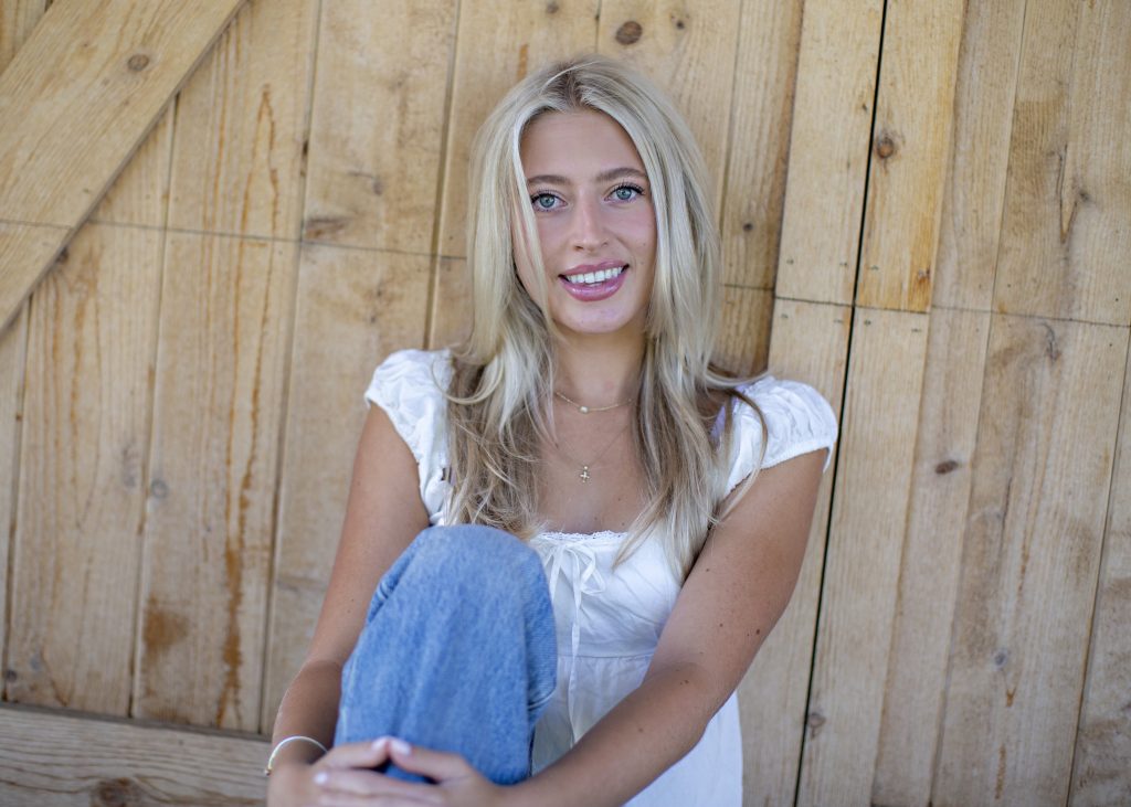 A smiling young woman with long blonde hair sits against a wooden wall, wearing a white top and blue jeans.