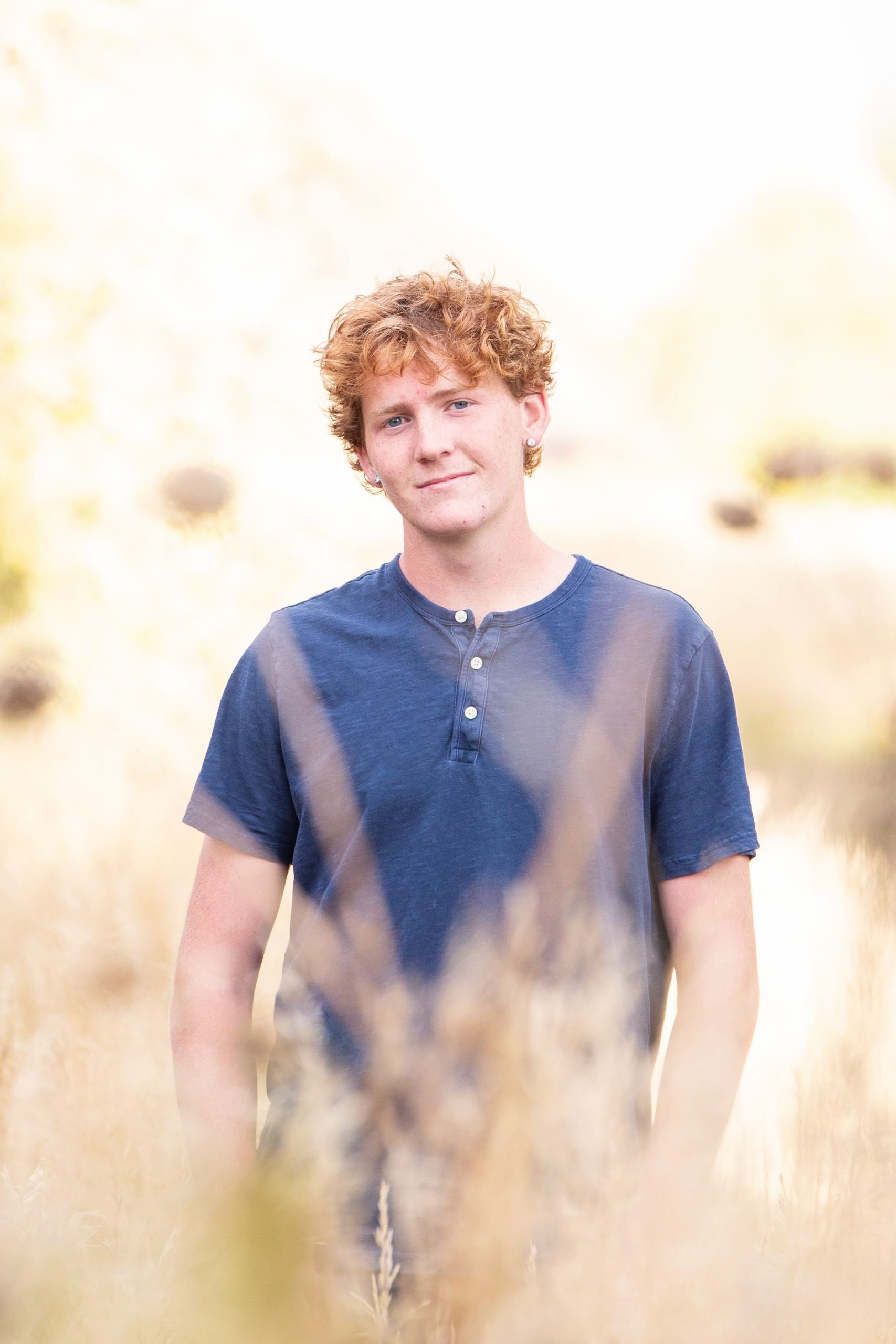 A young man with curly reddish hair stands among tall grasses, smiling softly at the camera in a natural setting.