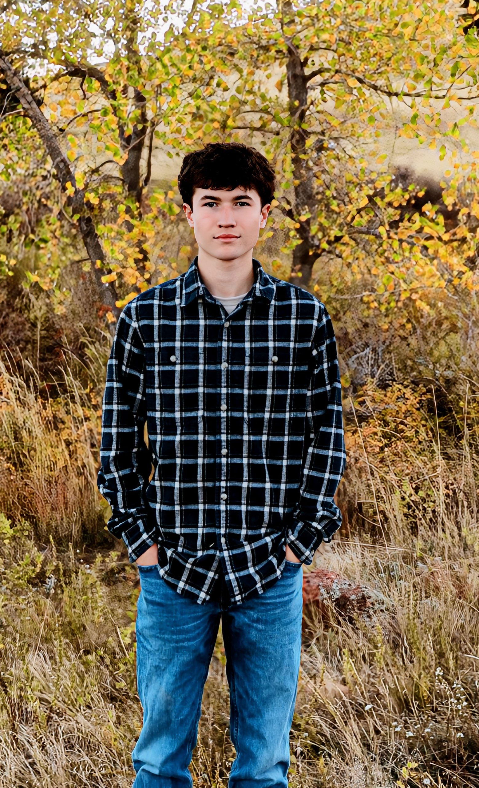 A young man stands in a field, wearing a black and white checkered shirt and blue jeans, with autumn trees in the background.