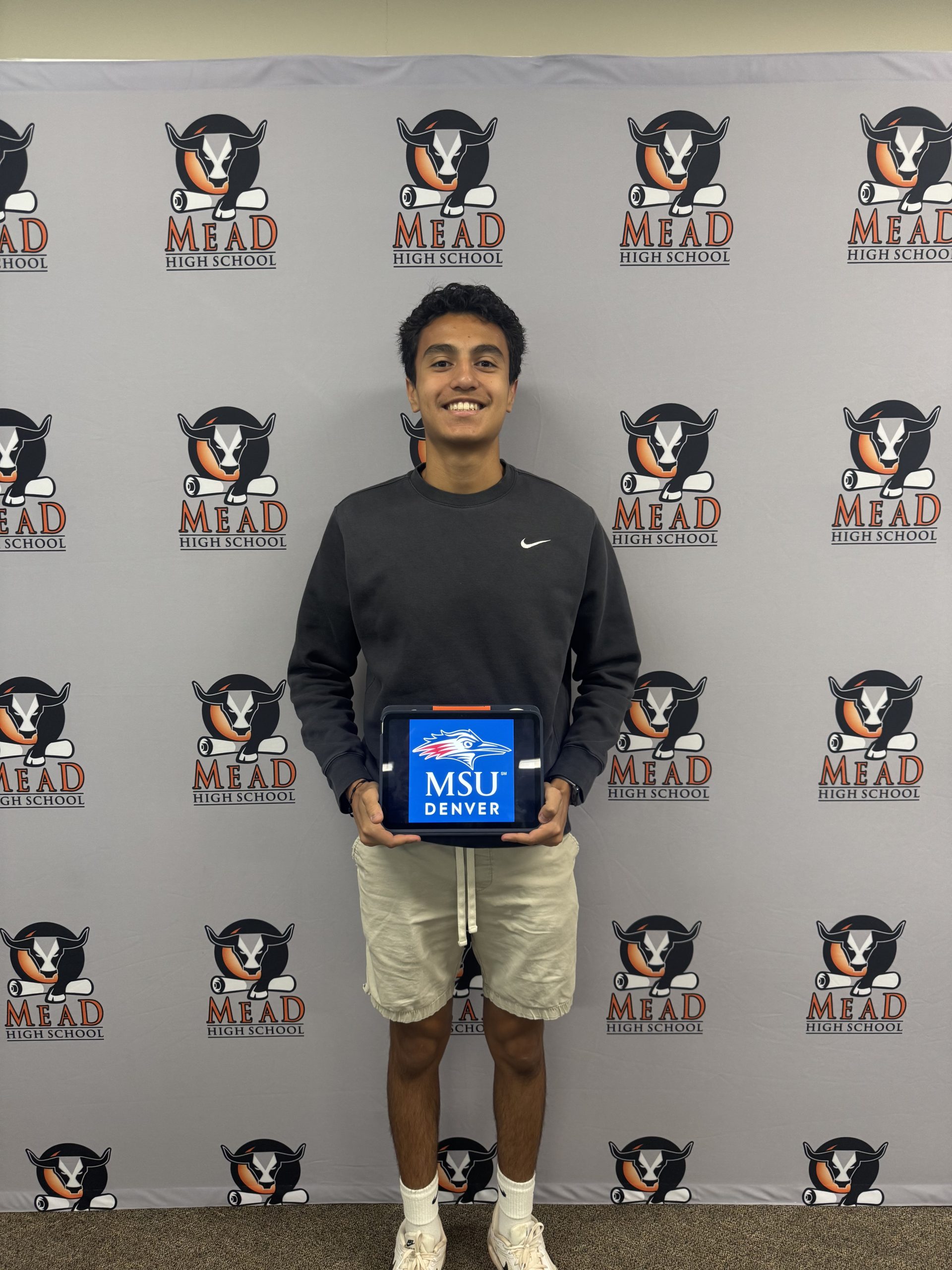 A student stands in front of a Mead High School backdrop, holding a tablet displaying the MSU Denver logo.