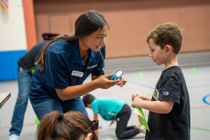 A young woman in a blue shirt interacts with a boy, showing him a robot, in a gym filled with children.