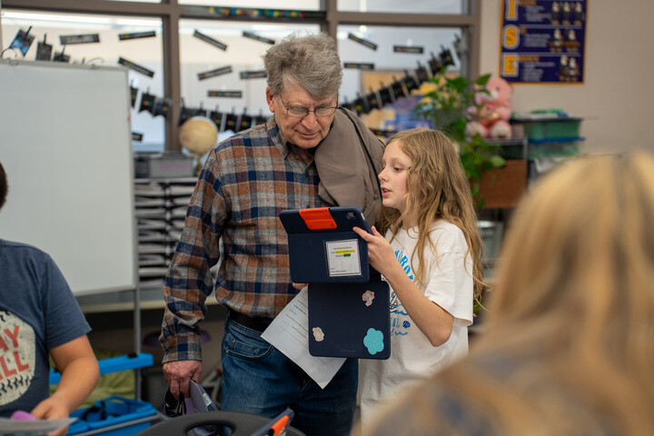 A girl shows a tablet to an older man in a classroom, while other students work at their desks in the background.