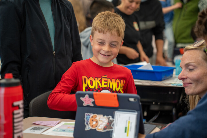 A smiling boy in a red Pok&eacute;mon shirt interacts with a tablet while an adult watches, surrounded by others.