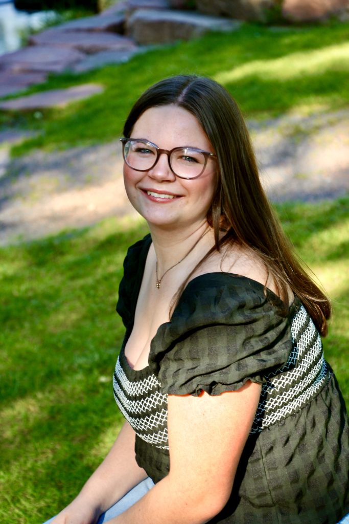 A young woman with long brown hair and glasses smiles while sitting on grass, wearing a black and white top.